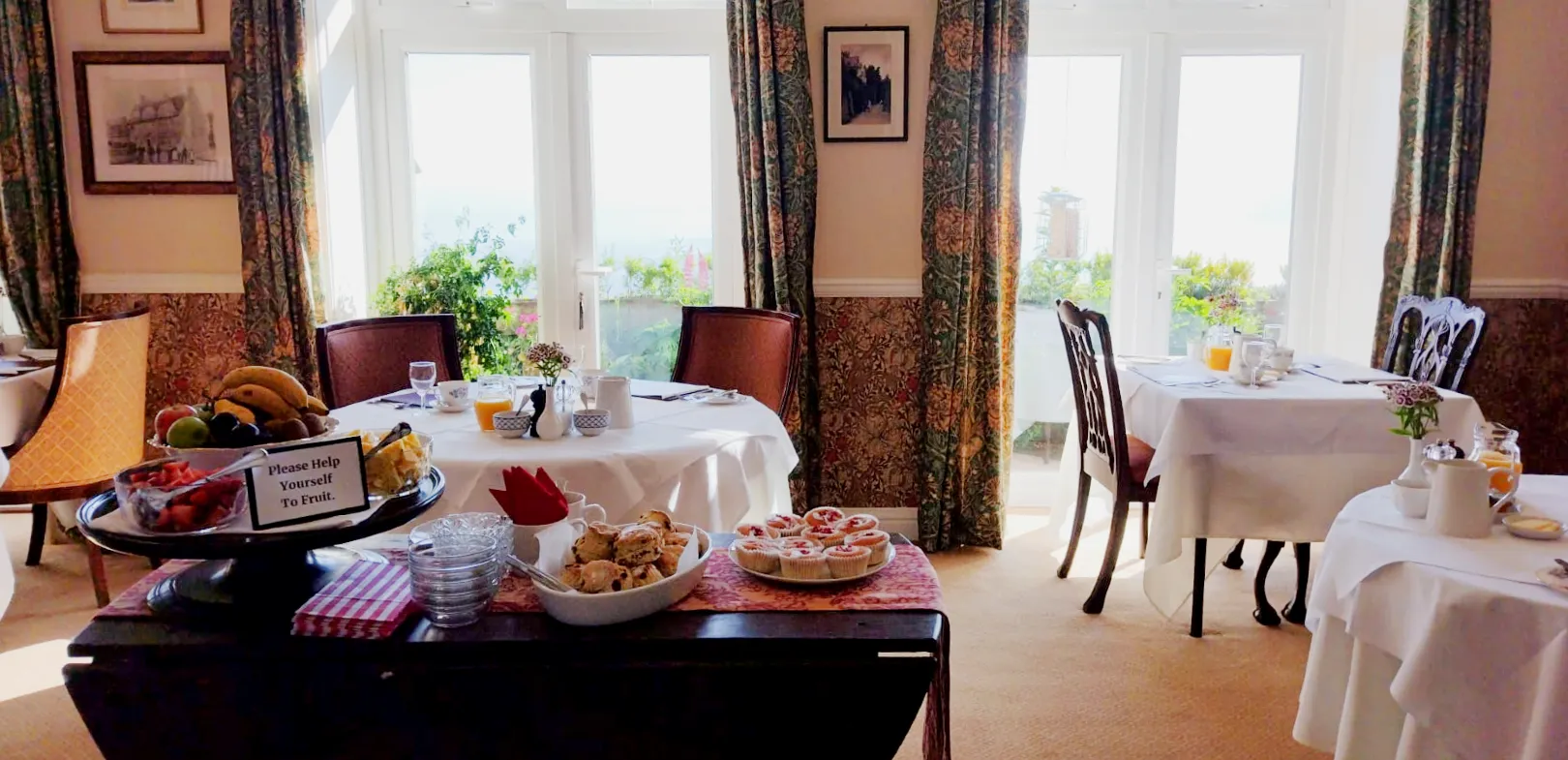 A dining room with tables set for breakfast, featuring a fruit platter, scones, and cupcakes on a side table