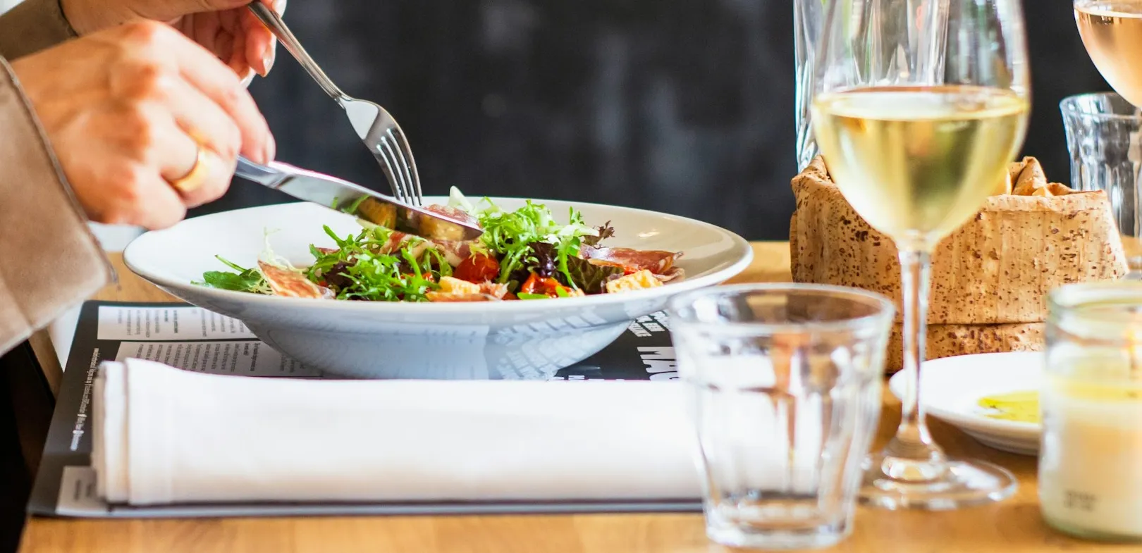 Person cutting into a fresh salad with a glass of white wine on a restaurant table