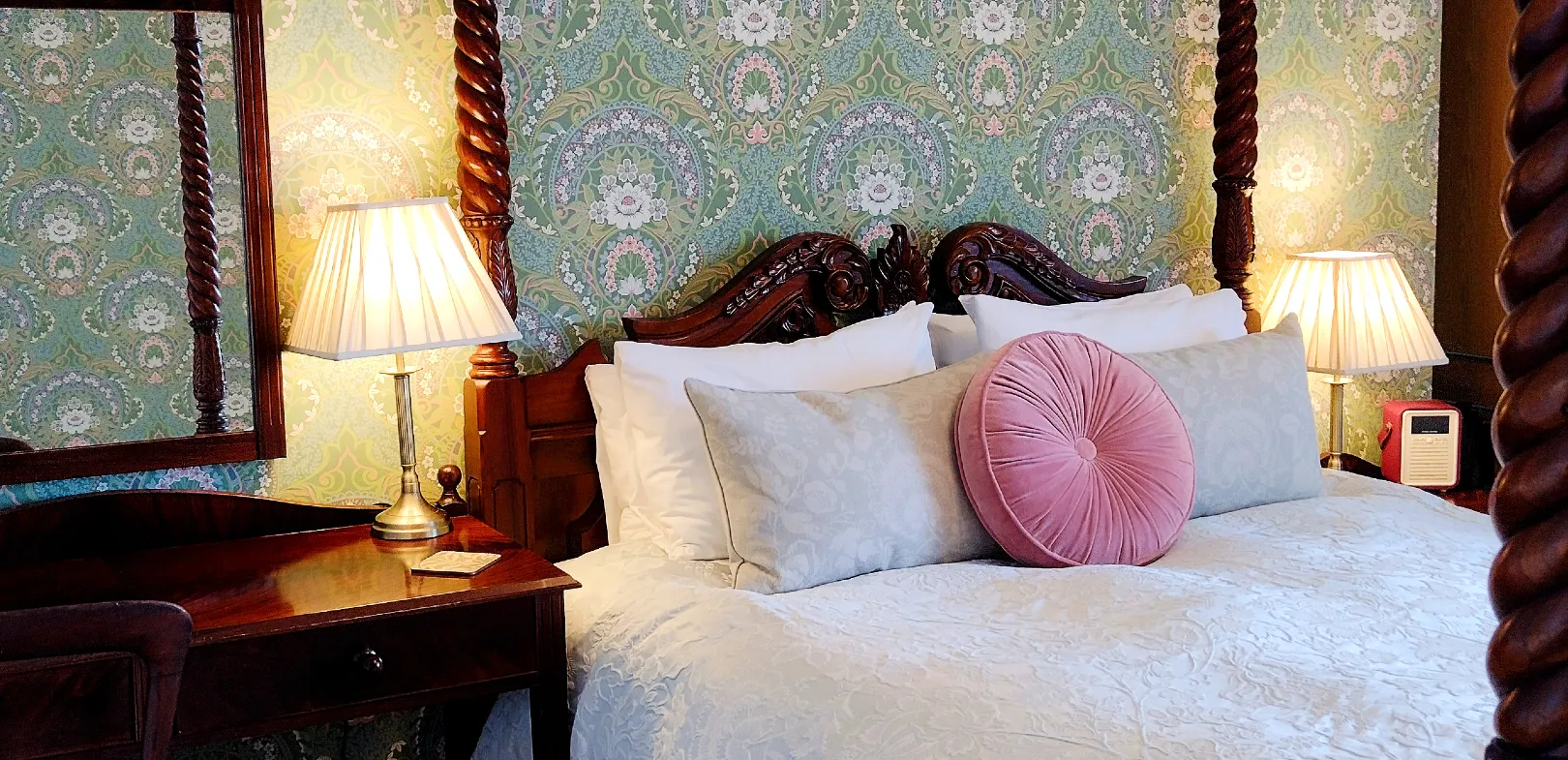 The 'Nest' four-poster room at Chough's Nest Hotel. Four-poster bed with carved wooden headboard, white bedding, decorative cushions, bedside tables with lamps, and floral wallpaper