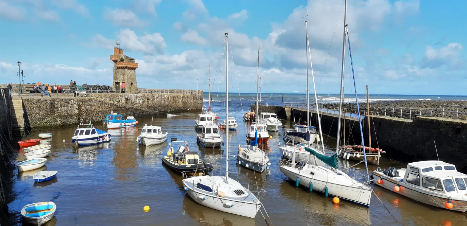 Small boats moored in Lynmouth Harbour with a stone pier and a tower under a partly cloudy sky