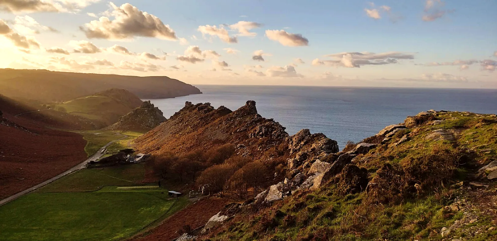 Valley of Rocks, Lynton. Coastal landscape with rocky hills, grassy fields, and the sea under a partly cloudy sky at sunset